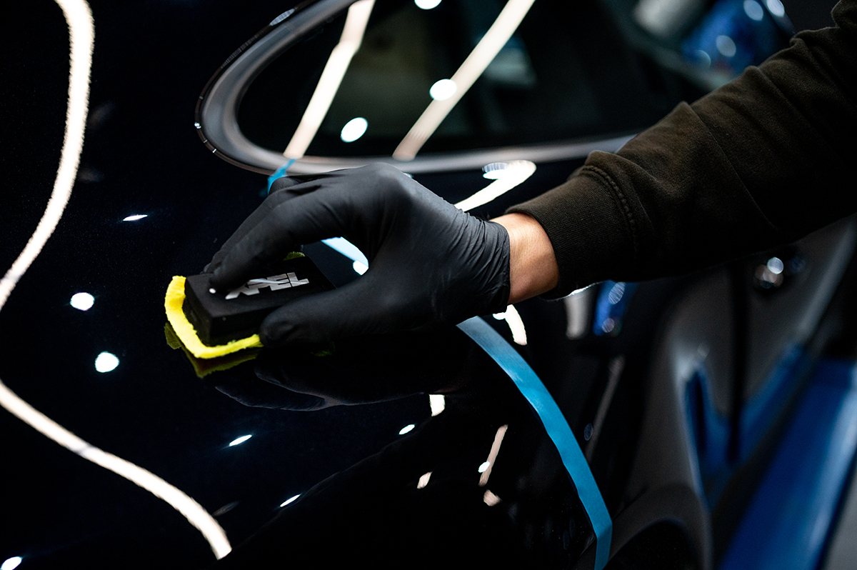 Detaljni pogled na majstora koji aplikuje zaštitni premaz na crnu automobilsku površinu koristeći žuti aplikator, sa neonom osvjetljenjem u pozadini